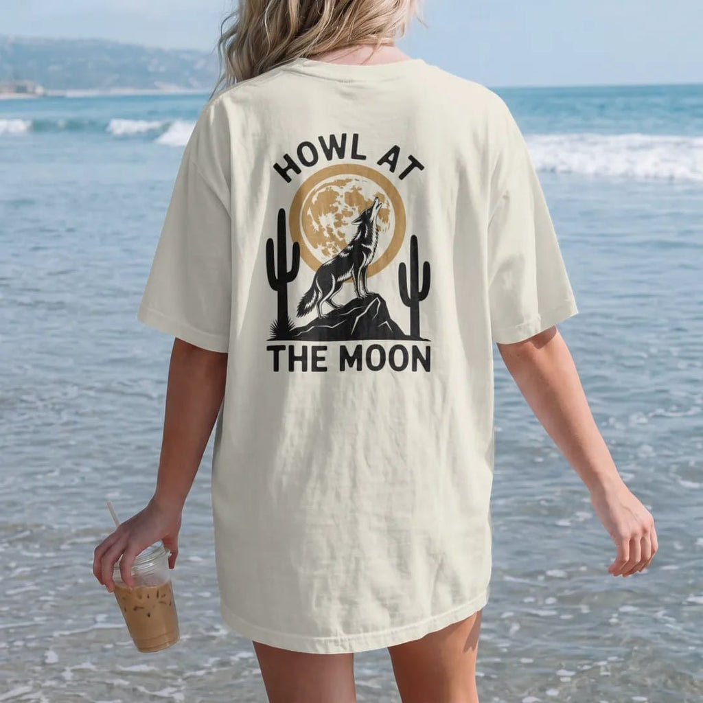 Woman on the beach in a Ivory tee, back view: a coyote on a rock howling at a big full moon with saguaros below, "HOWL AT" in small retro 70s caps above and "THE MOON" below, in a faded 1970s souvenir style with a worn cracked-ink texture, printed…