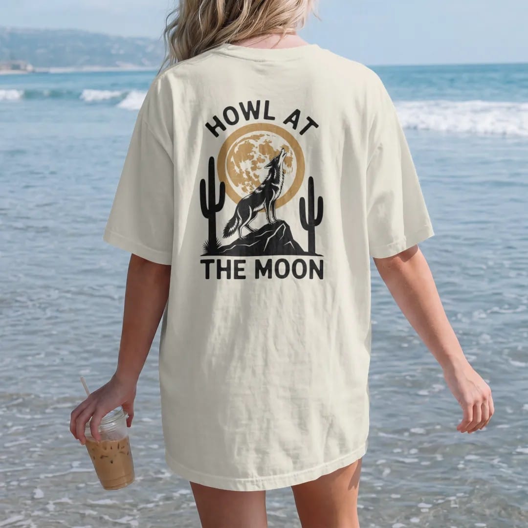 Woman on the beach in a Ivory tee, back view: a coyote on a rock howling at a big full moon with saguaros below, "HOWL AT" in small retro 70s caps above and "THE MOON" below, in a faded 1970s souvenir style with a worn cracked-ink texture, printed…