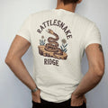 Man wearing a Ivory tee, seen from the back: a coiled rattlesnake on a rocky ledge with a few desert flowers, "RATTLESNAKE" in a faded arched Tuscan rodeo-poster header and "RIDGE" small below, in a faded 1970s souvenir style with a worn…
