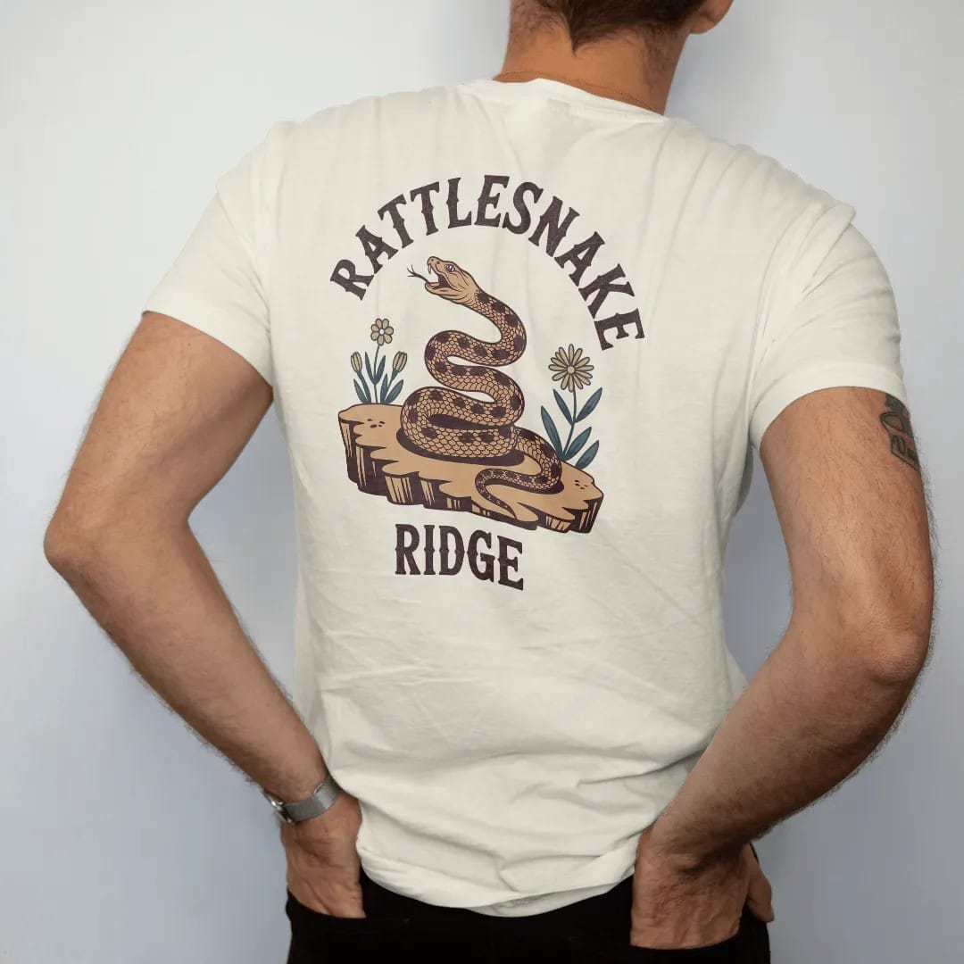Man wearing a Ivory tee, seen from the back: a coiled rattlesnake on a rocky ledge with a few desert flowers, "RATTLESNAKE" in a faded arched Tuscan rodeo-poster header and "RIDGE" small below, in a faded 1970s souvenir style with a worn…