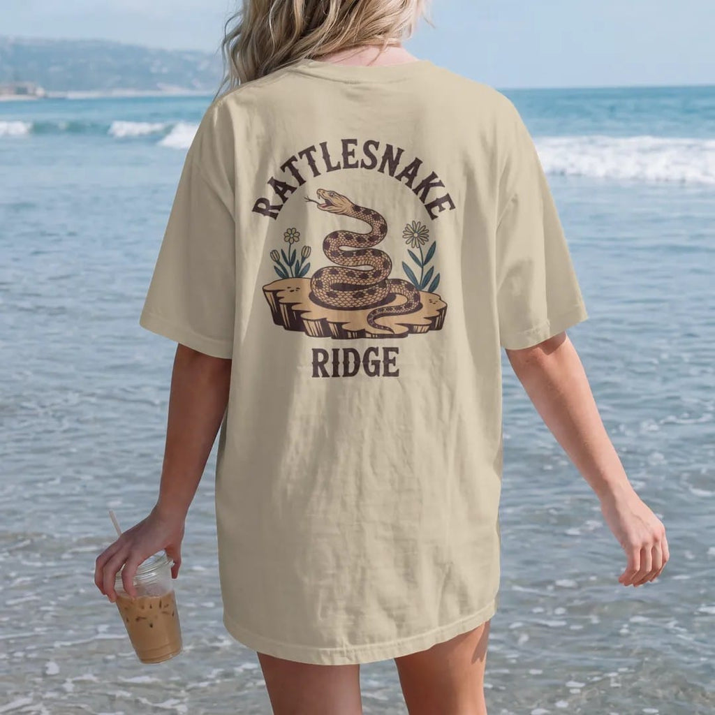 Woman on the beach in a Sandstone tee, back view: a coiled rattlesnake on a rocky ledge with a few desert flowers, "RATTLESNAKE" in a faded arched Tuscan rodeo-poster header and "RIDGE" small below, in a faded 1970s souvenir style with a worn…