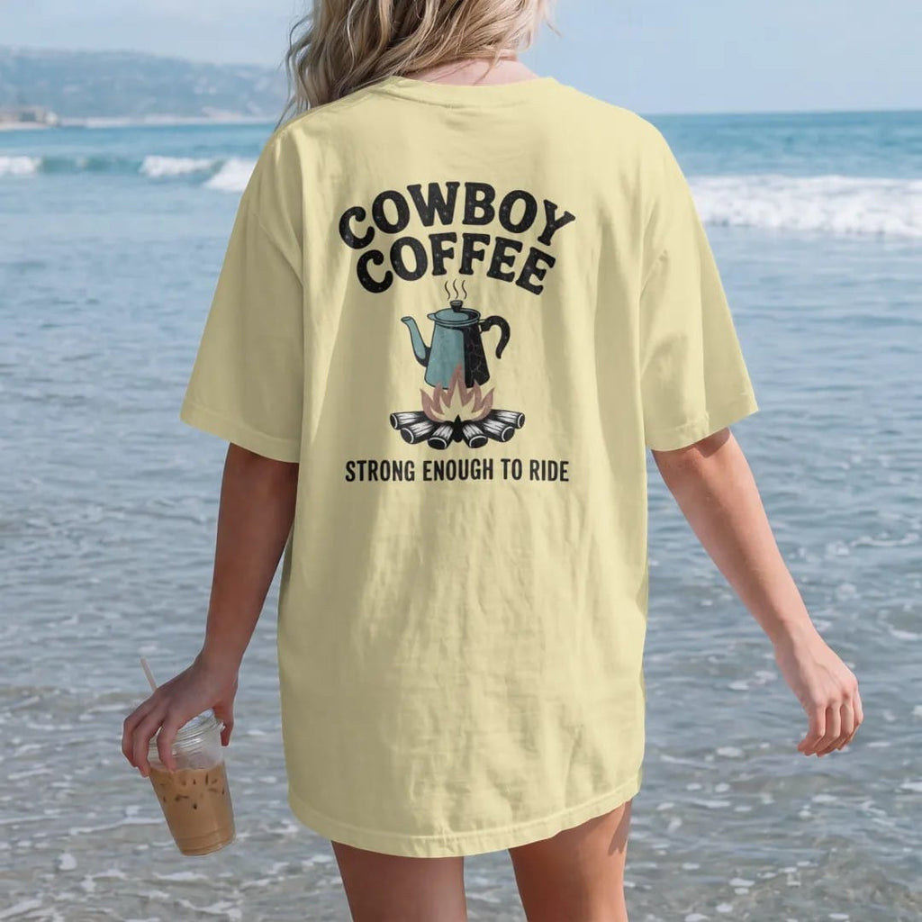 Woman on the beach in a Butter tee, back view: vintage ad parody of an enamel coffee pot over a campfire with steam, "COWBOY COFFEE" in a banner of chunky rounded 70s letters and "STRONG ENOUGH TO RIDE" small below, in a faded 1970s souvenir style…