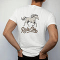 Man wearing a White tee, seen from the back: a galloping horse with flowing mane in mid-stride, "RIDE ON" in a looping 70s rope script beneath, in a faded 1970s souvenir style with a worn cracked-ink texture, printed in faded bone, sunset orange,…
