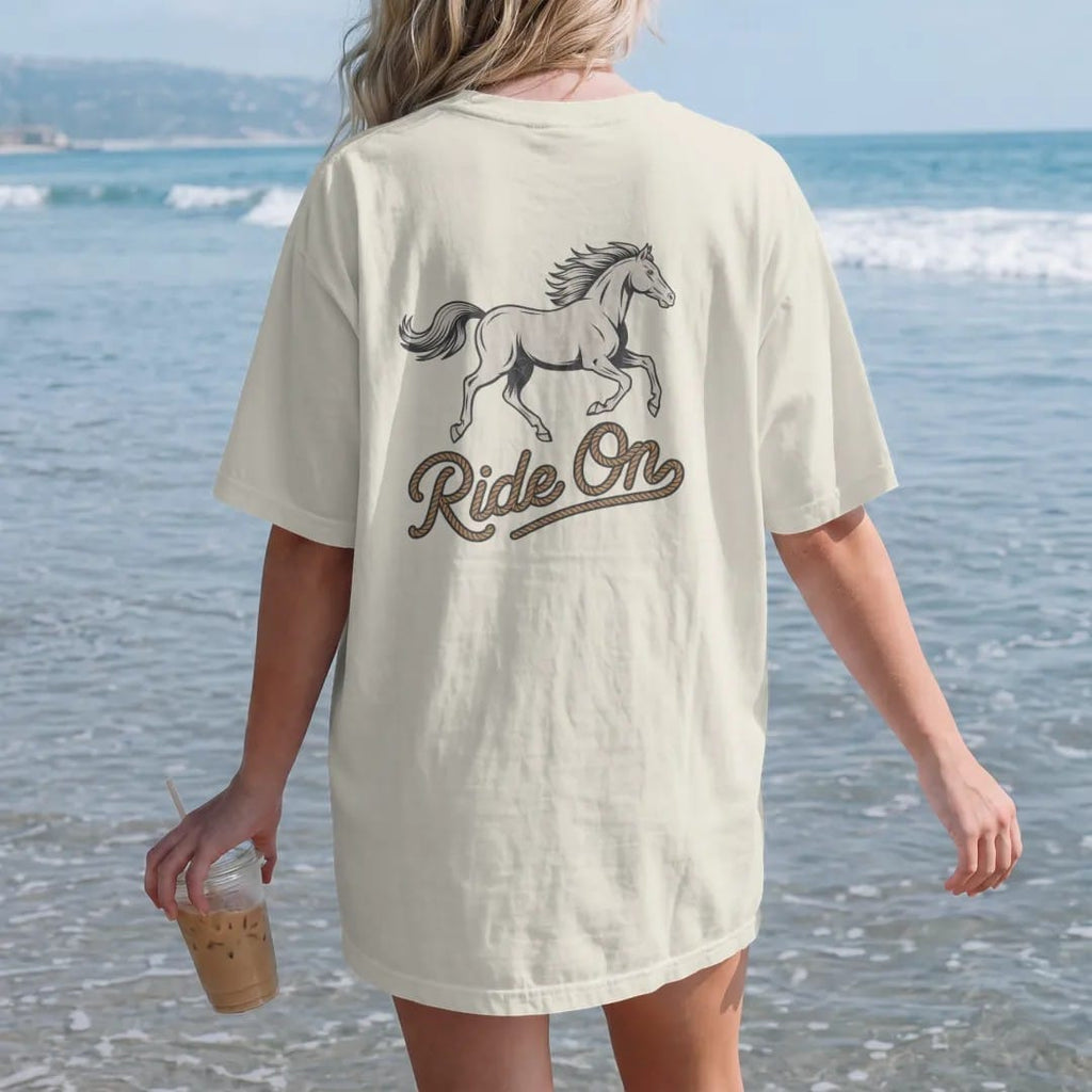 Woman on the beach in a Ivory tee, back view: a galloping horse with flowing mane in mid-stride, "RIDE ON" in a looping 70s rope script beneath, in a faded 1970s souvenir style with a worn cracked-ink texture, printed in faded bone, sunset orange,…