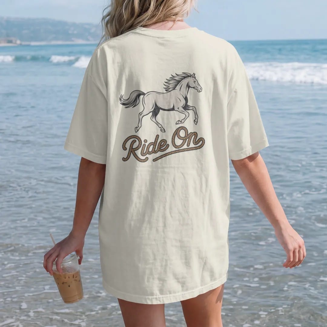Woman on the beach in a Ivory tee, back view: a galloping horse with flowing mane in mid-stride, "RIDE ON" in a looping 70s rope script beneath, in a faded 1970s souvenir style with a worn cracked-ink texture, printed in faded bone, sunset orange,…