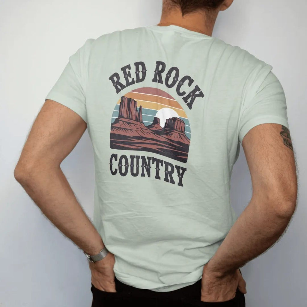Man wearing a Bay tee, seen from the back: layered red rock buttes and mesas under a sliced setting sun, "RED ROCK" in chunky 70s slab letters above and "COUNTRY" below, in a faded 1970s souvenir style with a worn cracked-ink texture, printed in…