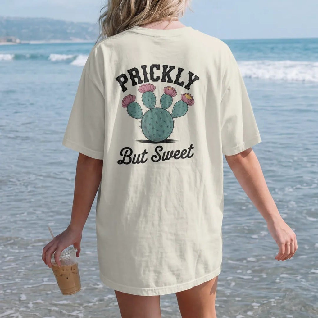 Woman on the beach in a Ivory tee, back view: a round prickly pear cactus with bright blooms and fruit, "PRICKLY" in chunky 70s block letters above and "BUT SWEET" in a looping 70s rope script below, in a faded 1970s souvenir style with a worn…
