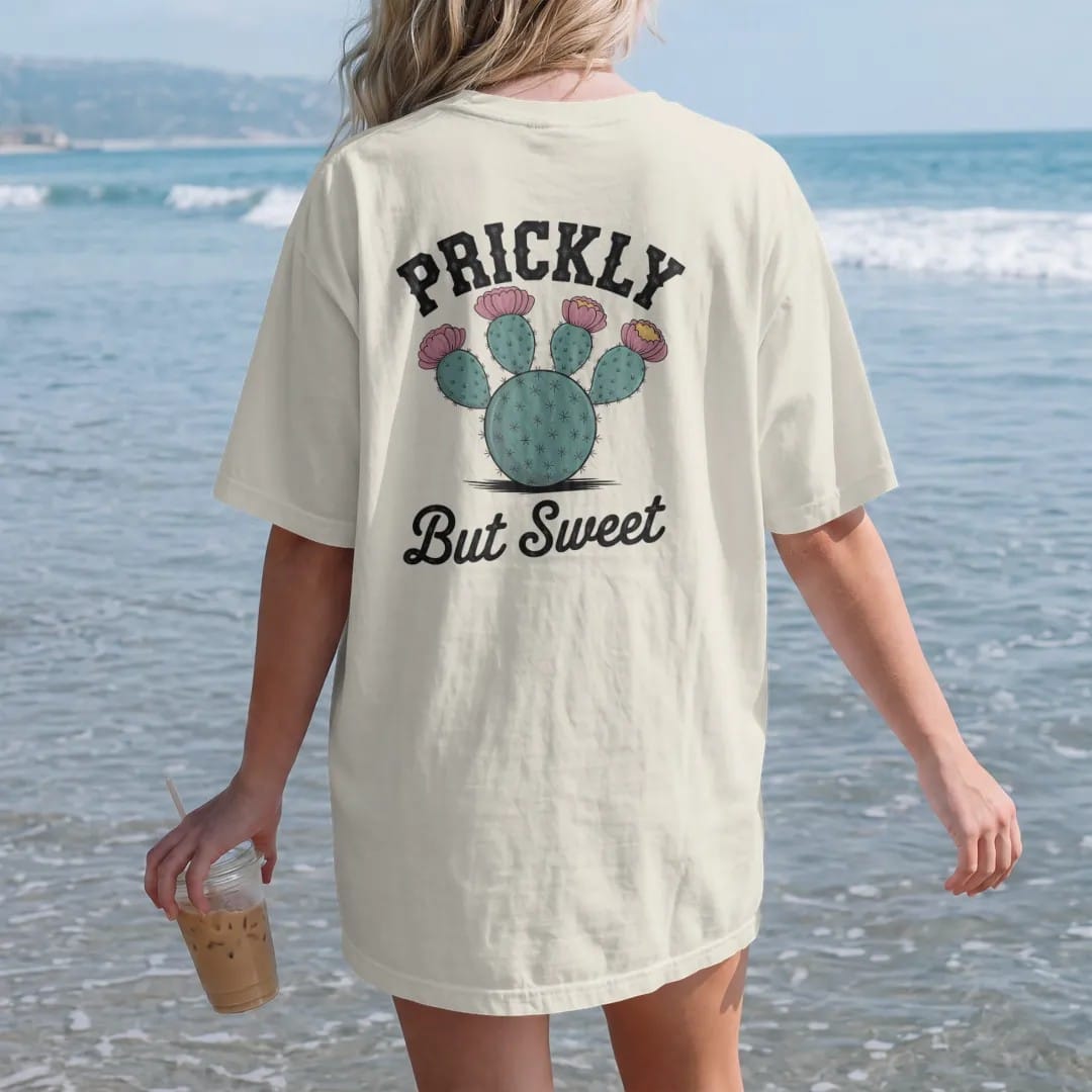 Woman on the beach in a Ivory tee, back view: a round prickly pear cactus with bright blooms and fruit, "PRICKLY" in chunky 70s block letters above and "BUT SWEET" in a looping 70s rope script below, in a faded 1970s souvenir style with a worn…
