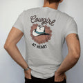 Man wearing a Granite tee, seen from the back: a cowgirl hat with a turquoise band resting on a heart-shaped wreath of desert flowers, "COWGIRL" in a looping 70s rope script above and "AT HEART" small below, in a faded 1970s souvenir style with a…