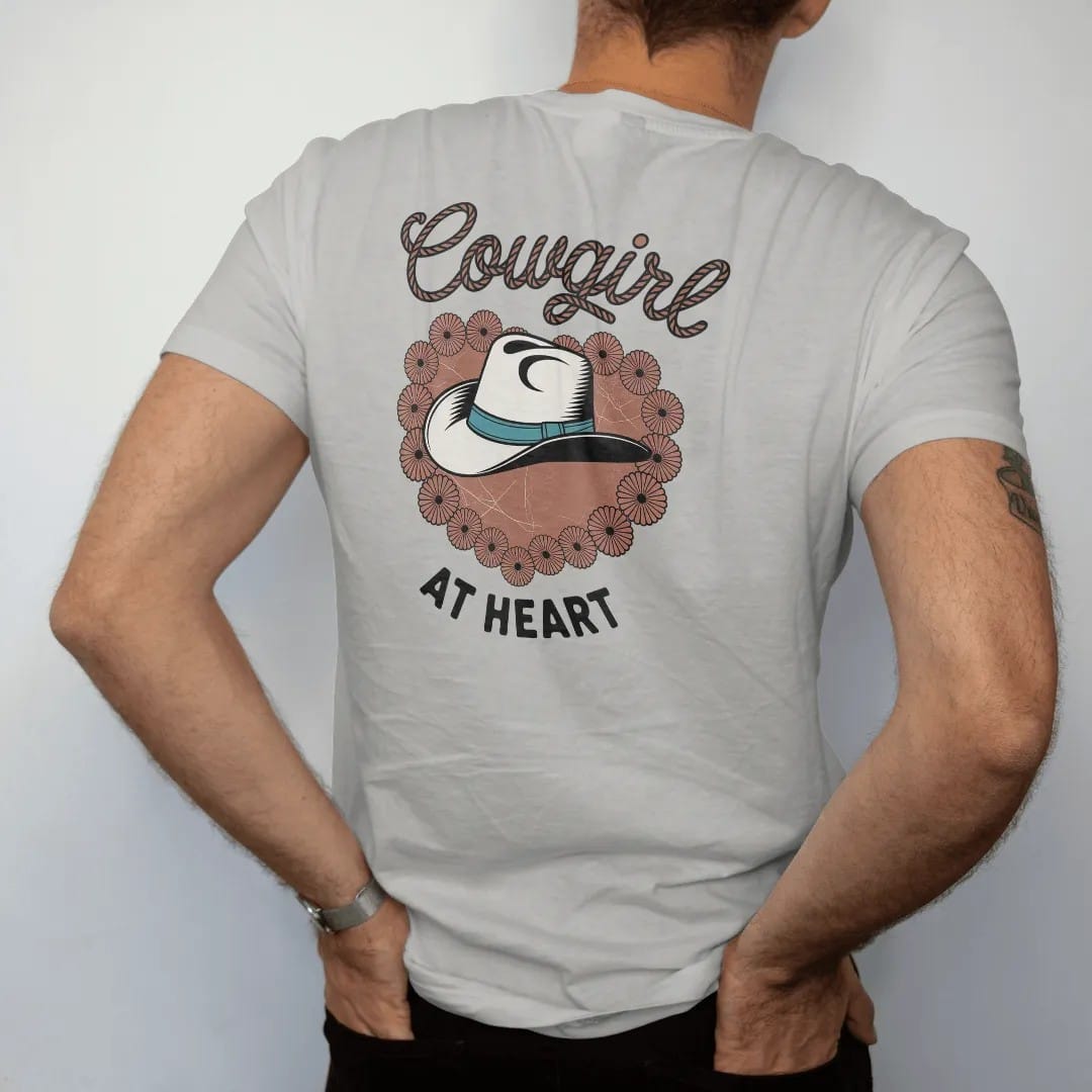 Man wearing a Granite tee, seen from the back: a cowgirl hat with a turquoise band resting on a heart-shaped wreath of desert flowers, "COWGIRL" in a looping 70s rope script above and "AT HEART" small below, in a faded 1970s souvenir style with a…