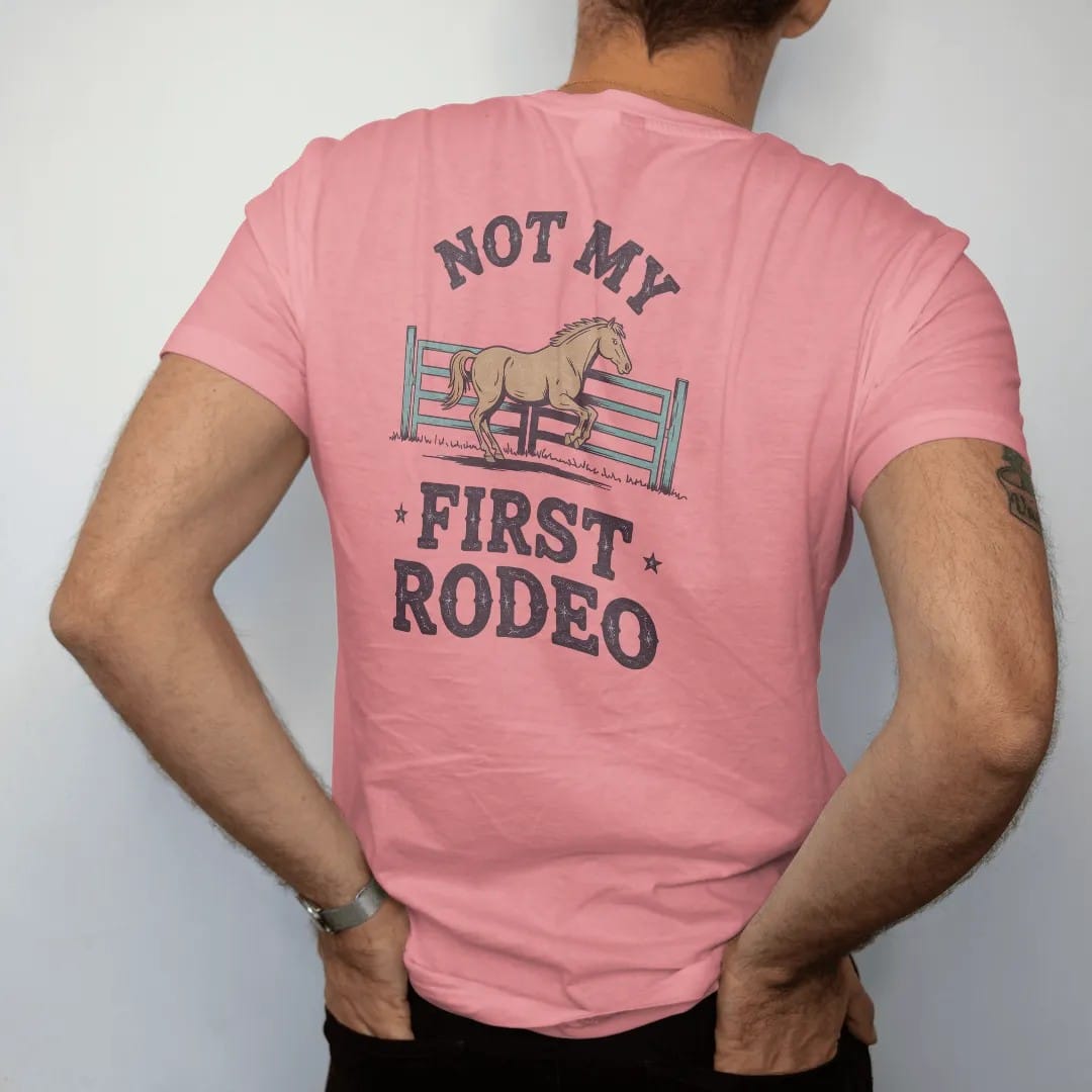 Man wearing a Watermelon tee, seen from the back: a dusty arena gate with a bronc kicking behind it, "NOT MY" in small retro 70s caps above and "FIRST RODEO" in chunky 70s slab letters below, in a faded 1970s souvenir style with a worn cracked-ink…