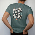 Man wearing a Blue Spruce tee, seen from the back: the single word "YEEHAW" in huge chunky 70s block letters with a small cowboy hat perched on the first Y and a few stars around it, in a faded 1970s souvenir style with a worn cracked-ink texture,…