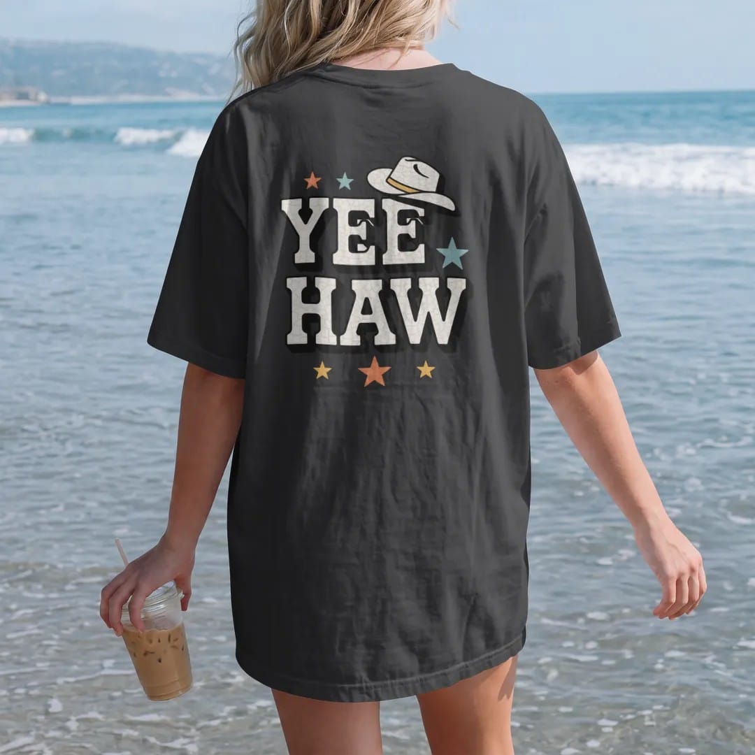 Woman on the beach in a Black tee, back view: the single word "YEEHAW" in huge chunky 70s block letters with a small cowboy hat perched on the first Y and a few stars around it, in a faded 1970s souvenir style with a worn cracked-ink texture,…