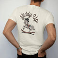 Man wearing a Ivory tee, seen from the back: a rocking horse with a cowboy hat, "GIDDY UP" in a looping 70s rope script arched above, in a faded 1970s souvenir style with a worn cracked-ink texture, printed in saddle brown, faded rust, dusty…