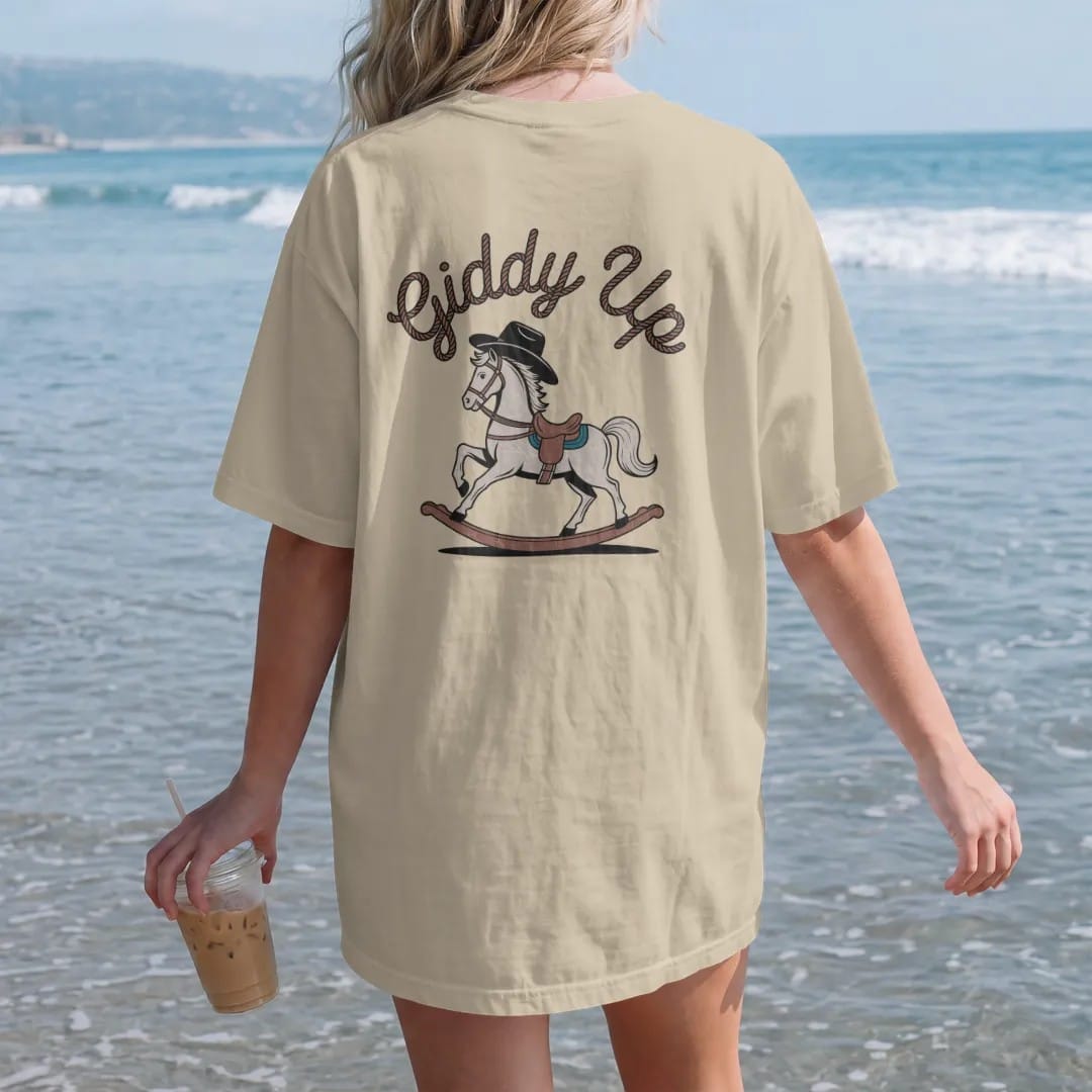 Woman on the beach in a Sandstone tee, back view: a rocking horse with a cowboy hat, "GIDDY UP" in a looping 70s rope script arched above, in a faded 1970s souvenir style with a worn cracked-ink texture, printed in saddle brown, faded rust, dusty…