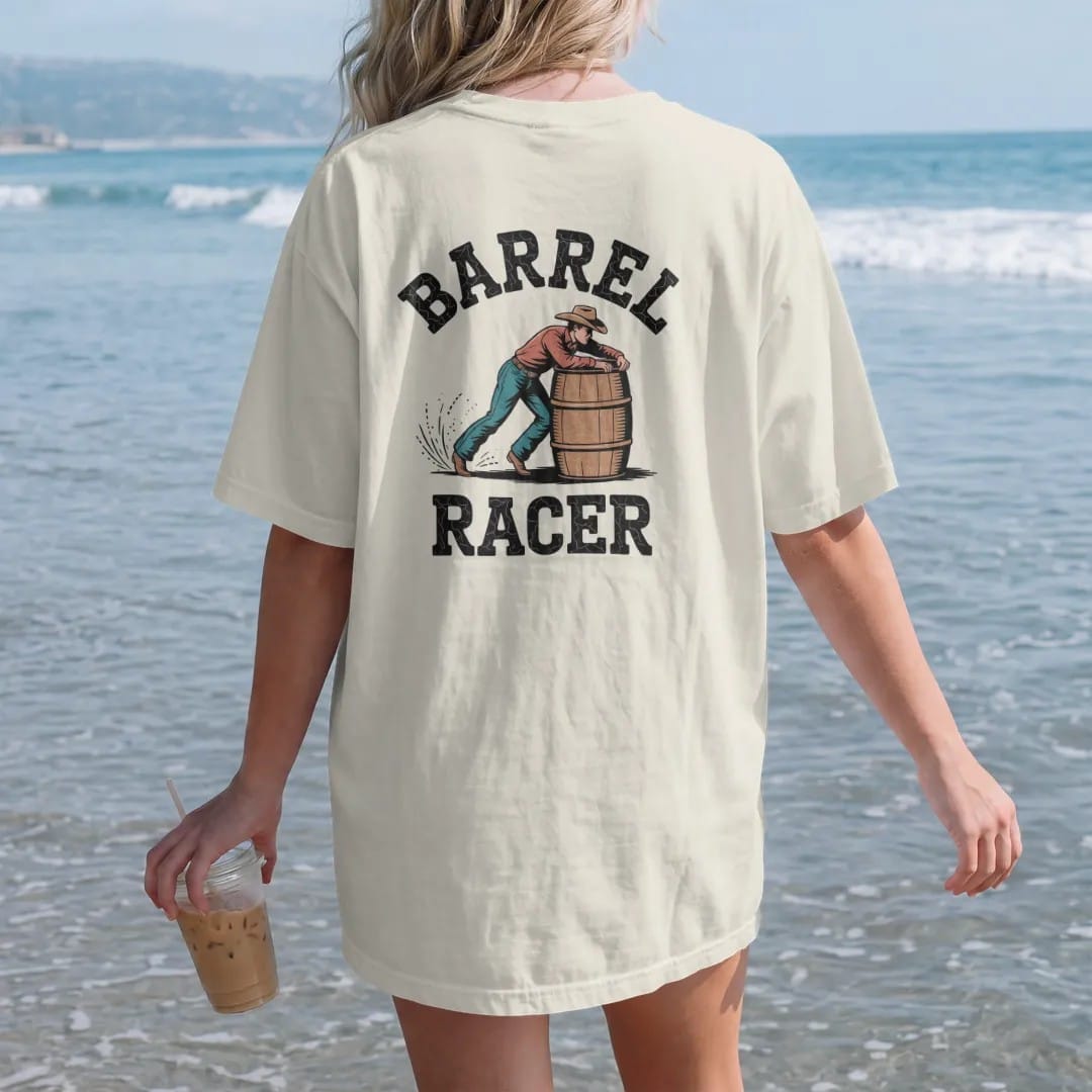Woman on the beach in a Ivory tee, back view: a barrel racer leaning hard around a barrel with dust flying, "BARREL" arched above and "RACER" below in chunky 70s slab letters, in a faded 1970s souvenir style with a worn cracked-ink texture, printed…