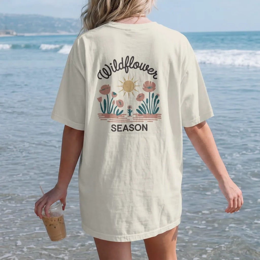 Woman on the beach in a Ivory tee, back view: desert floor in bloom with poppies, lupines and a small cactus flower under a soft sun, "WILDFLOWER" in a looping 70s rope script above and "SEASON" below, in a faded 1970s souvenir style with a worn…