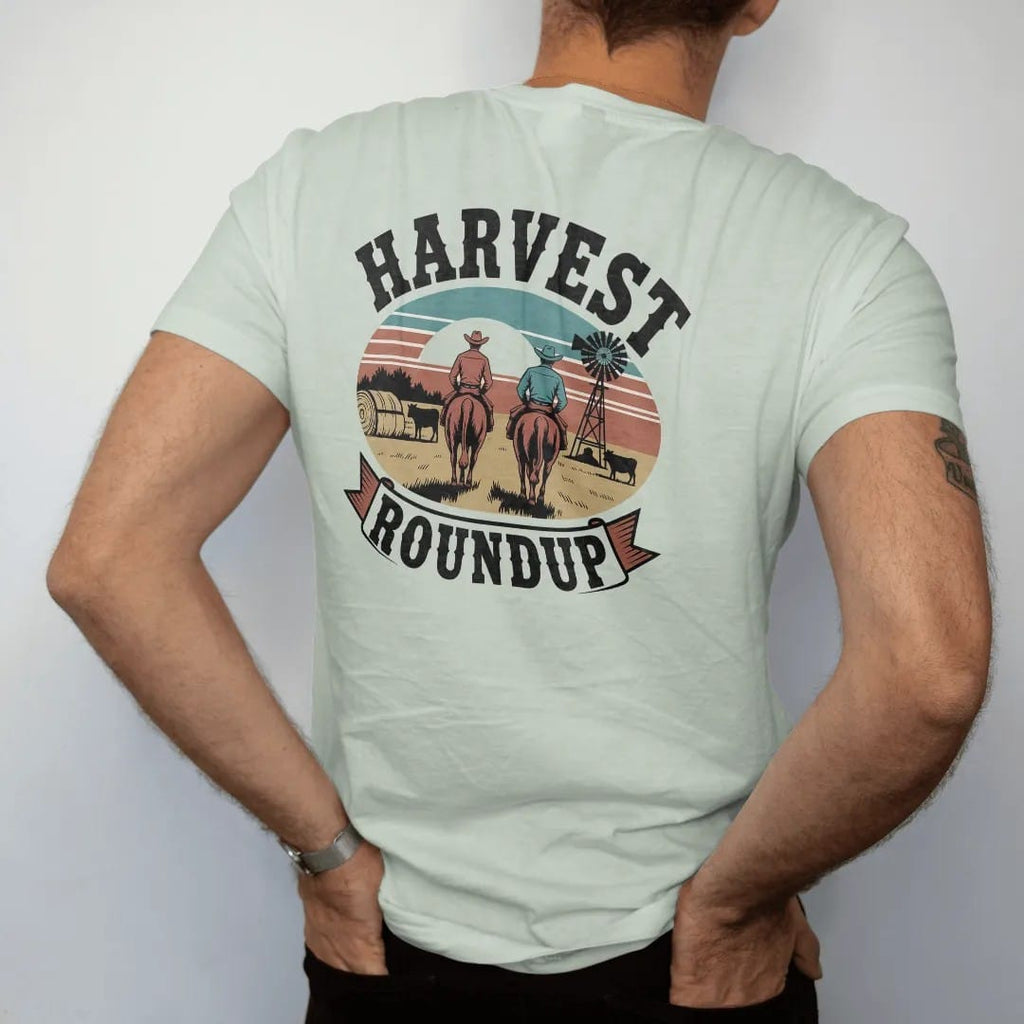 Man wearing a Bay tee, seen from the back: two cowboys driving cattle past hay bales and a windmill at golden hour, "HARVEST" in chunky 70s slab letters above and "ROUNDUP" on a banner below, in a faded 1970s souvenir style with a worn cracked-ink…