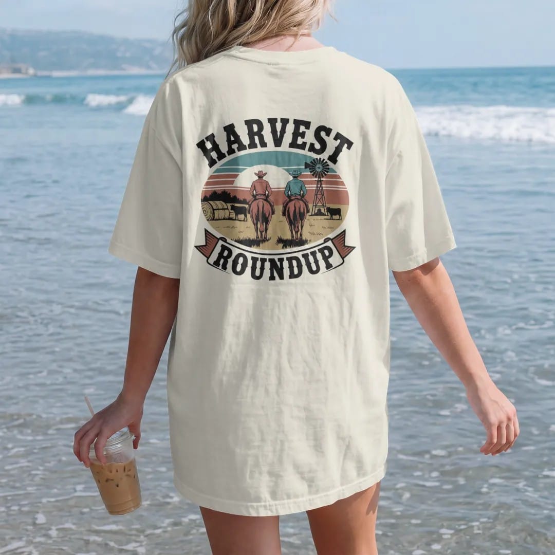 Woman on the beach in a Ivory tee, back view: two cowboys driving cattle past hay bales and a windmill at golden hour, "HARVEST" in chunky 70s slab letters above and "ROUNDUP" on a banner below, in a faded 1970s souvenir style with a worn…