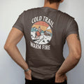 Man wearing a Espresso tee, seen from the back: a horse and rider in a wool coat on a snowy trail toward a small cabin with smoke rising, "COLD TRAIL" above and "WARM FIRE" below in small retro 70s caps, in a faded 1970s souvenir style with a worn…