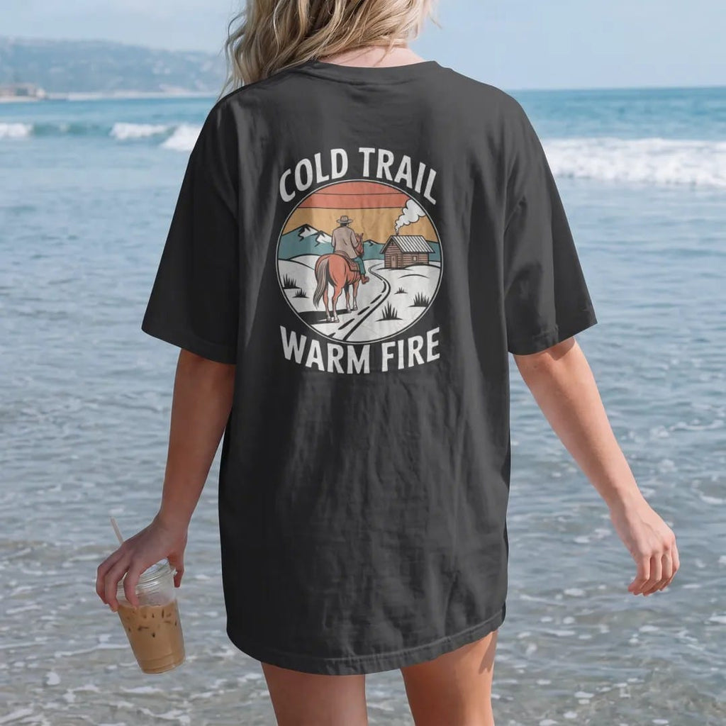 Woman on the beach in a Black tee, back view: a horse and rider in a wool coat on a snowy trail toward a small cabin with smoke rising, "COLD TRAIL" above and "WARM FIRE" below in small retro 70s caps, in a faded 1970s souvenir style with a worn…