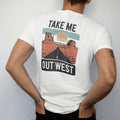 Man wearing a White tee, seen from the back: a vintage travel postcard scene of mesas, a winding road and a big sun, "TAKE ME" in small caps above and "OUT WEST" in large faded 70s souvenir-sign lettering below, in a faded 1970s souvenir style with…