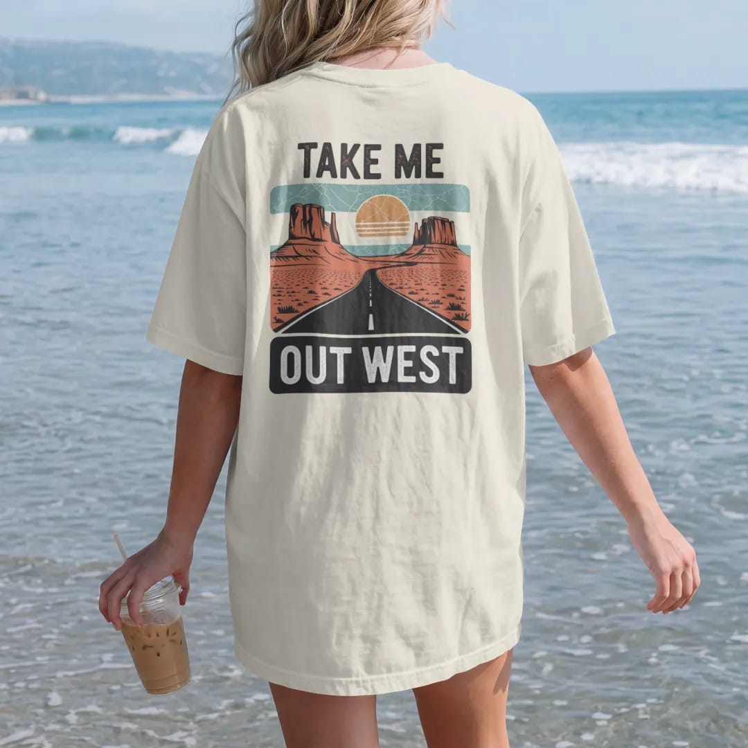 Woman on the beach in a Ivory tee, back view: a vintage travel postcard scene of mesas, a winding road and a big sun, "TAKE ME" in small caps above and "OUT WEST" in large faded 70s souvenir-sign lettering below, in a faded 1970s souvenir style…