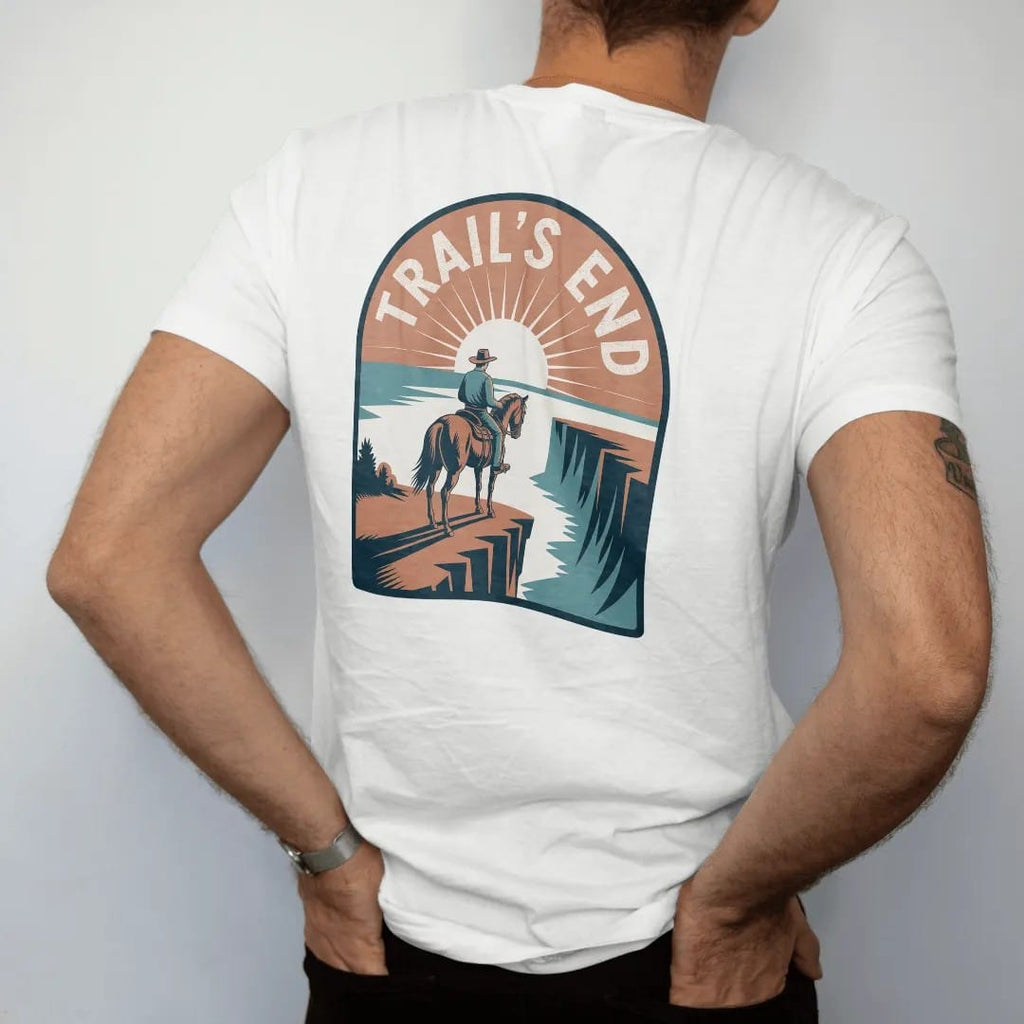 Man wearing a White tee, seen from the back: a tired horse and rider at the edge of a cliff overlooking a sunset valley, "TRAIL'S END" in a faded arched Tuscan rodeo-poster header above, in a faded 1970s souvenir style with a worn cracked-ink…
