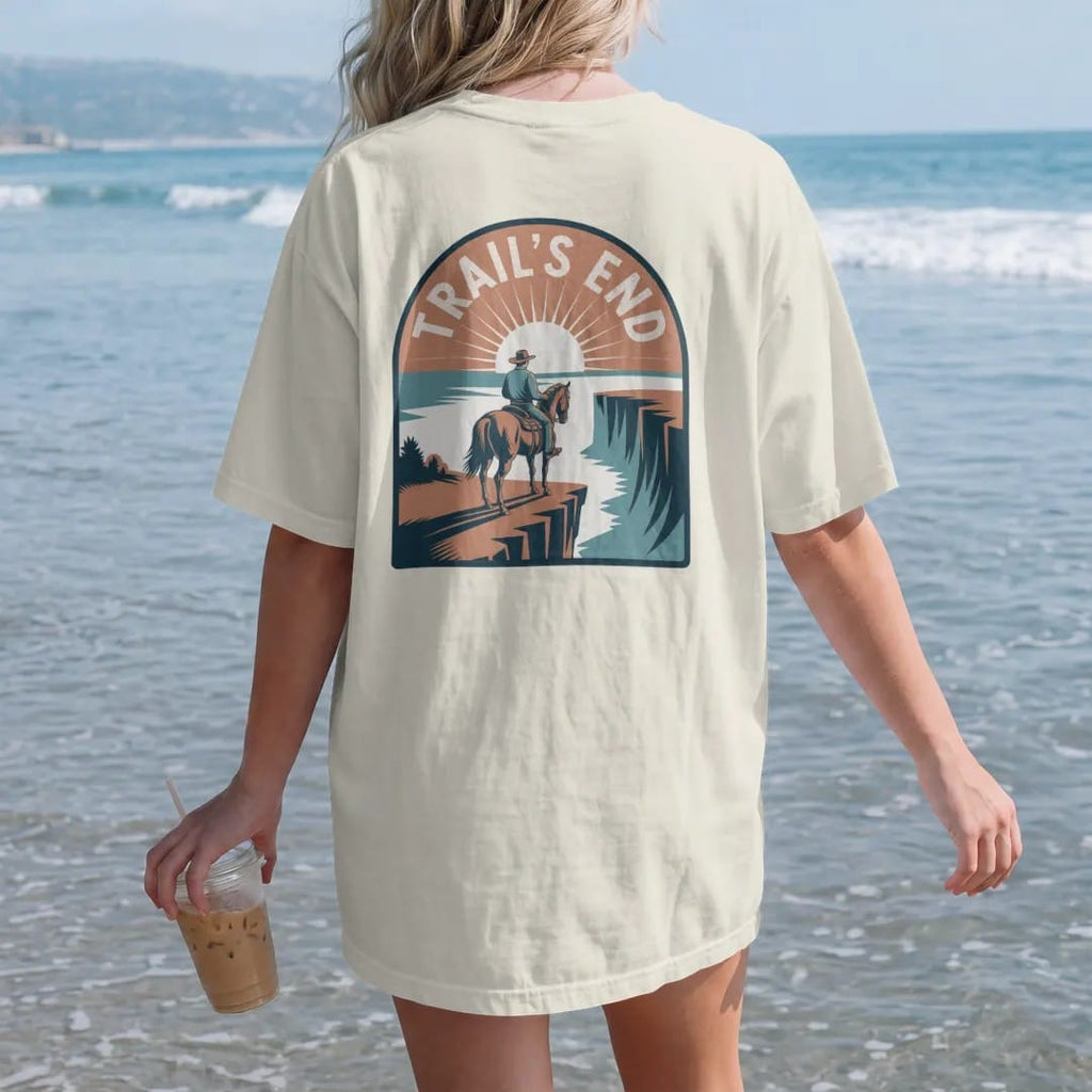 Woman on the beach in a Ivory tee, back view: a tired horse and rider at the edge of a cliff overlooking a sunset valley, "TRAIL'S END" in a faded arched Tuscan rodeo-poster header above, in a faded 1970s souvenir style with a worn cracked-ink…