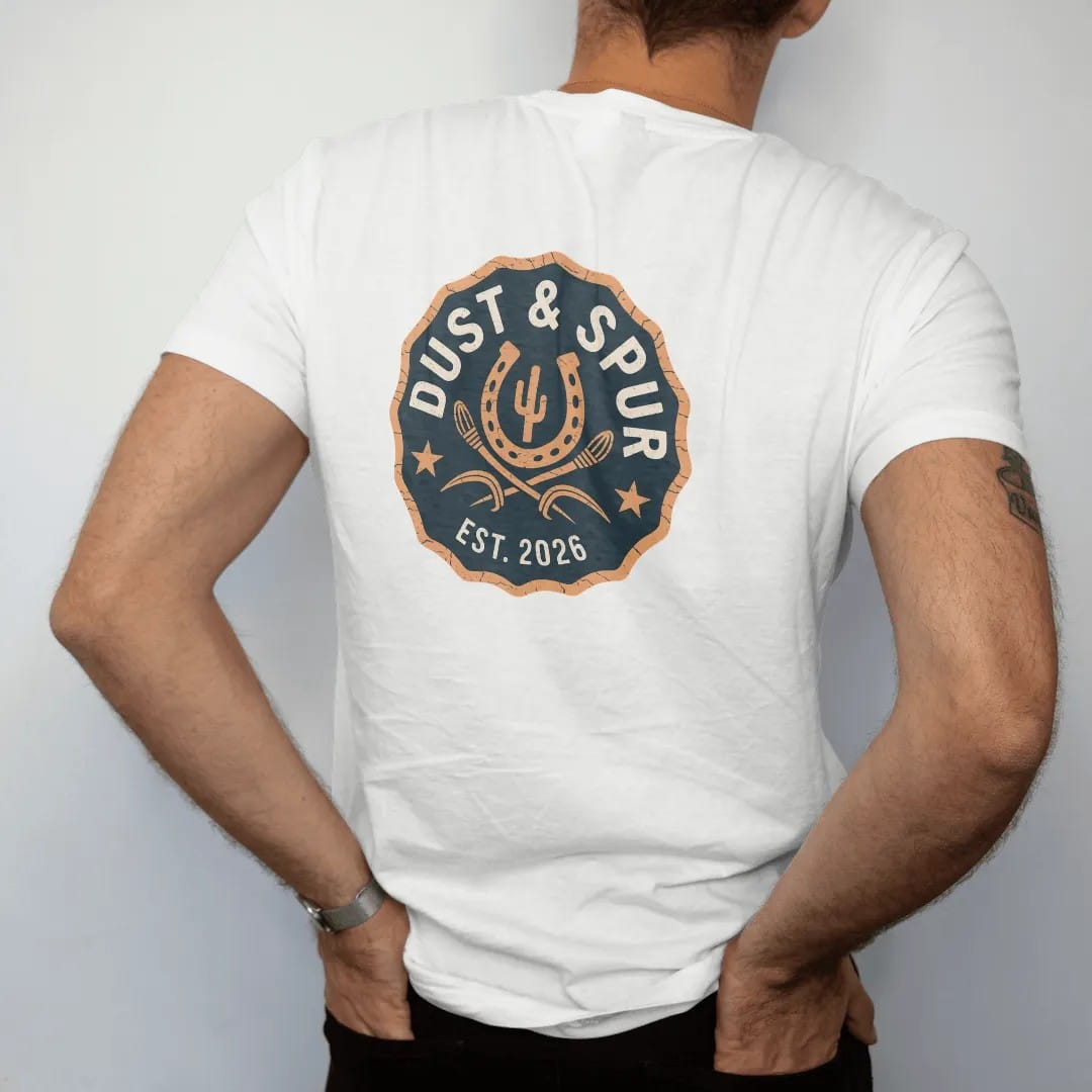 Man wearing a White tee, seen from the back: a round brand-style seal with a horseshoe, crossed spurs and a small cactus, "DUST & SPUR" arched in chunky 70s slab letters and "EST. 2026" below, in a faded 1970s souvenir style with a worn cracked-ink…