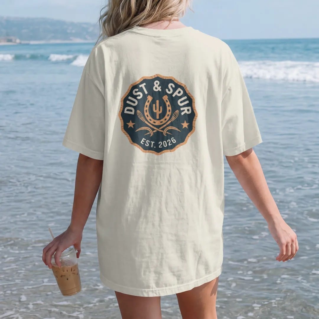 Woman on the beach in a Ivory tee, back view: a round brand-style seal with a horseshoe, crossed spurs and a small cactus, "DUST & SPUR" arched in chunky 70s slab letters and "EST. 2026" below, in a faded 1970s souvenir style with a worn…