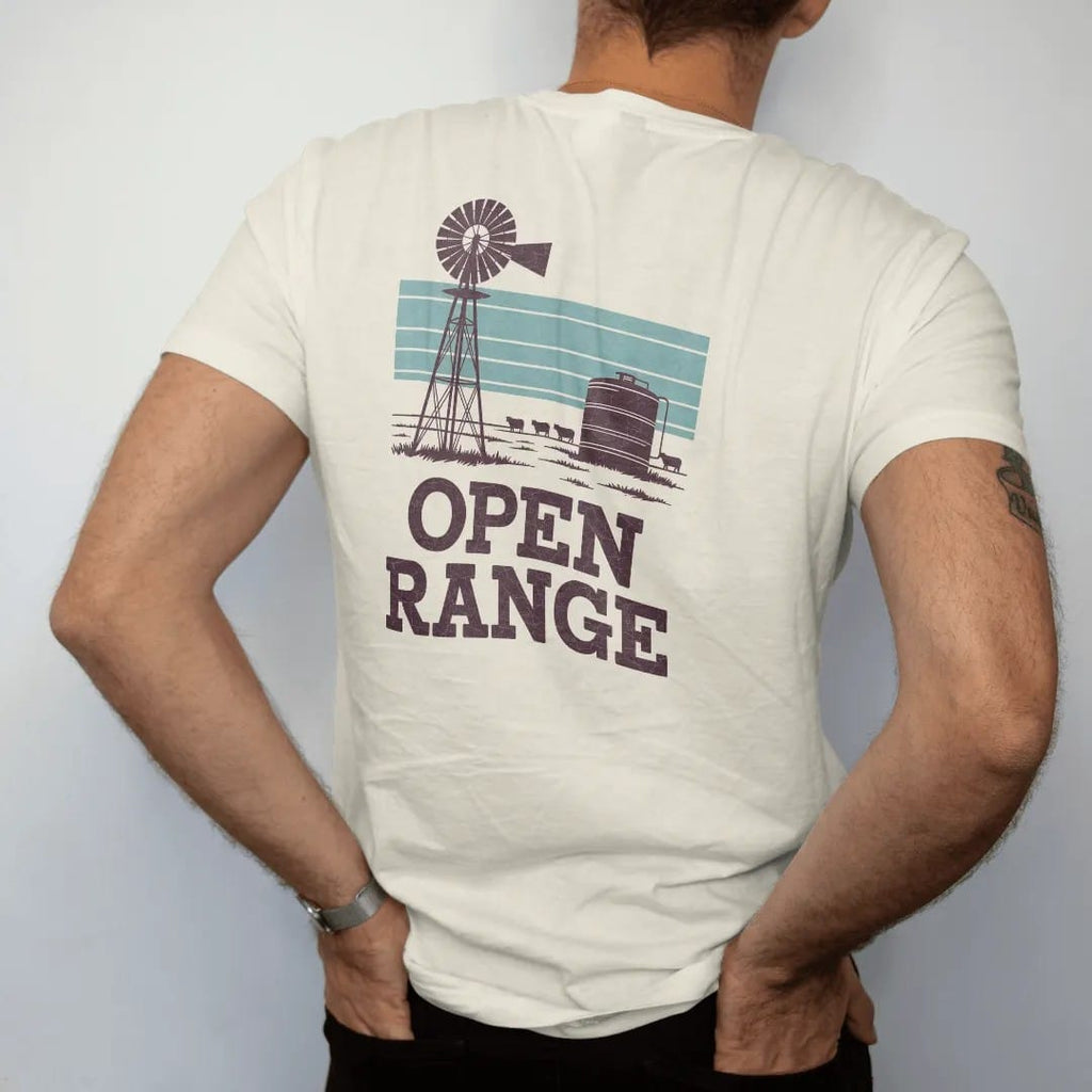 Man wearing a Ivory tee, seen from the back: an old ranch windmill and water tank on flat grassland under a wide sky with a few cattle, "OPEN RANGE" in chunky 70s slab letters beneath, in a faded 1970s souvenir style with a worn cracked-ink…
