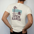 Man wearing a Ivory tee, seen from the back: an old ranch windmill and water tank on flat grassland under a wide sky with a few cattle, "OPEN RANGE" in chunky 70s slab letters beneath, in a faded 1970s souvenir style with a worn cracked-ink…