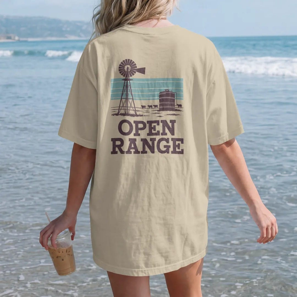 Woman on the beach in a Sandstone tee, back view: an old ranch windmill and water tank on flat grassland under a wide sky with a few cattle, "OPEN RANGE" in chunky 70s slab letters beneath, in a faded 1970s souvenir style with a worn cracked-ink…
