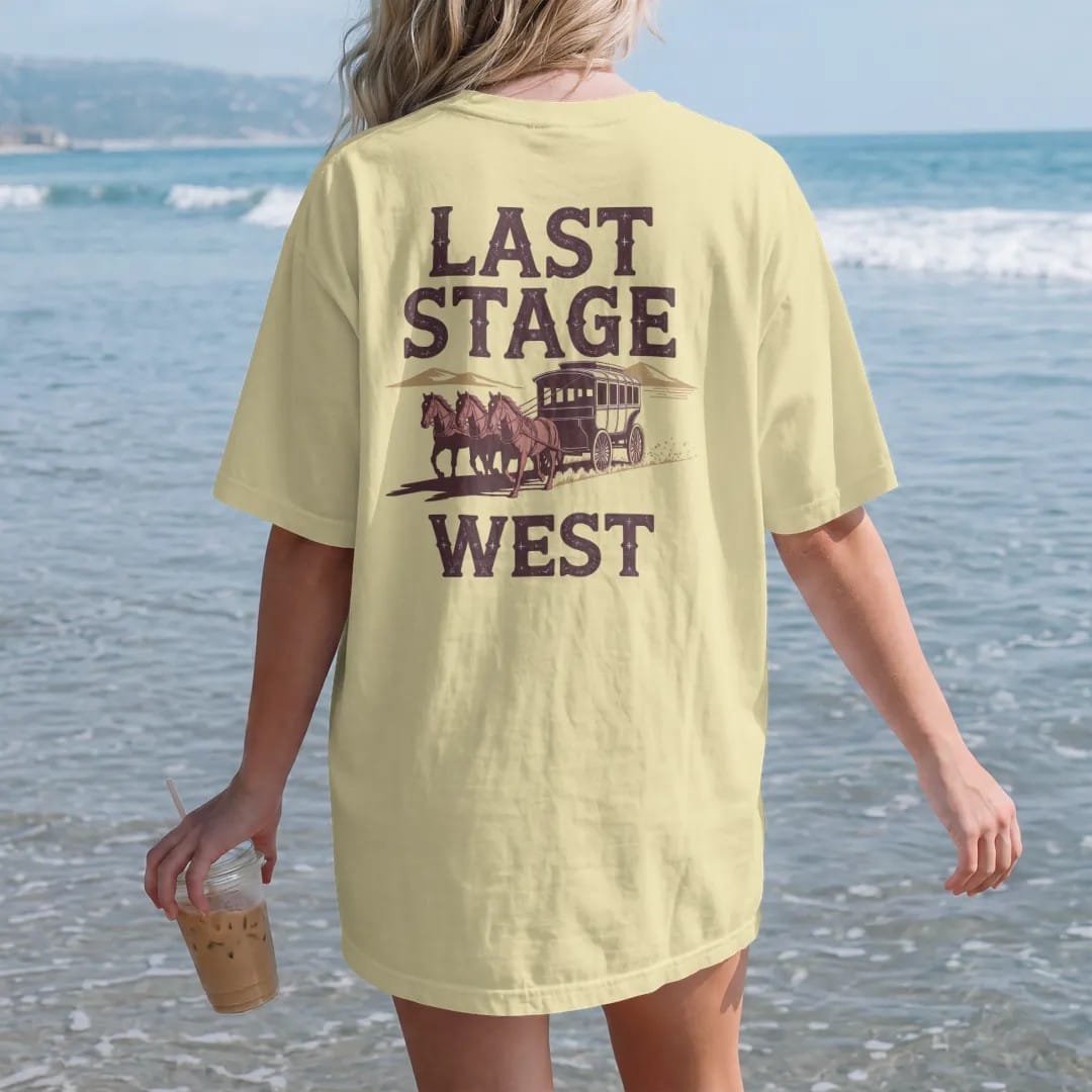 Woman on the beach in a Butter tee, back view: a stagecoach pulled by four horses across the desert with dust trailing behind, "LAST STAGE" in faded Tuscan rodeo-poster letters above and "WEST" small below, in a faded 1970s souvenir style with a…