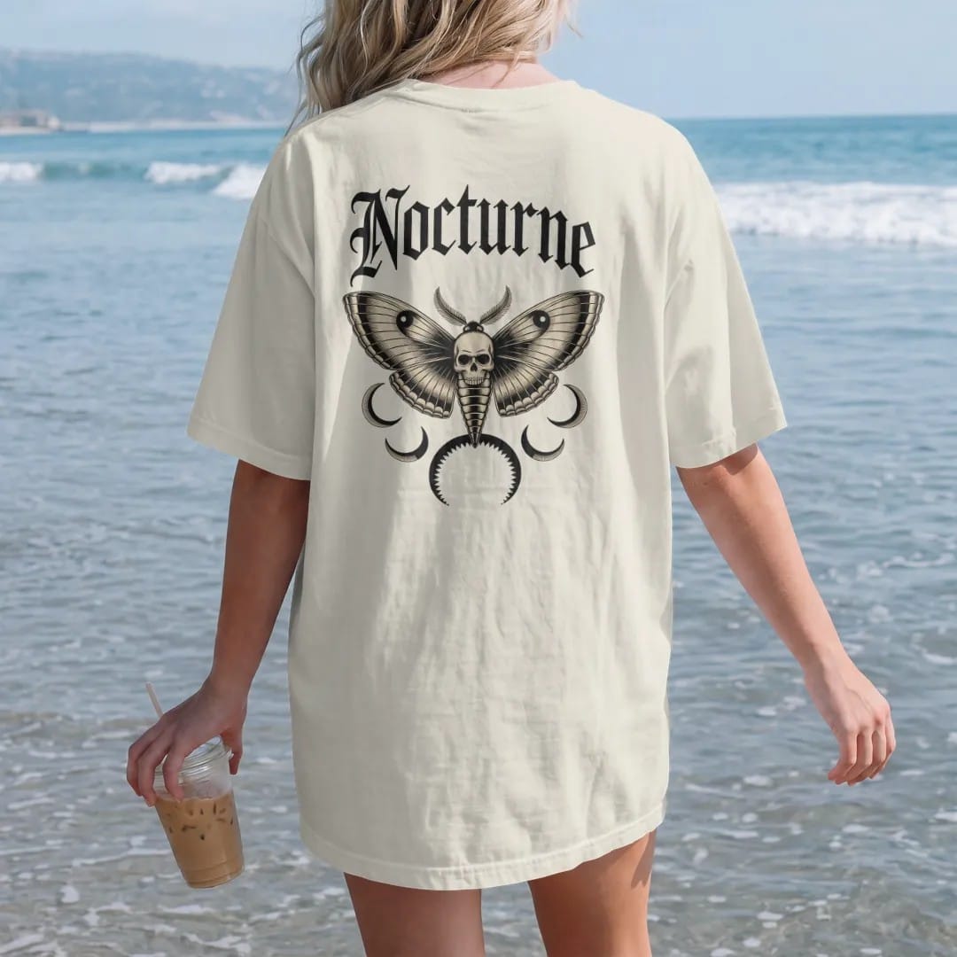 Woman on the beach in a Ivory tee, back view: back print: death's-head hawkmoth with open wings and a skull marking on its back, crescent moons around it, detailed Victorian engraving with crosshatching, "NOCTURNE" arched above in ornate gothic…