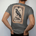 Man wearing a Pepper tee, seen from the back: back print: raven perched on a skull inside an ornate tarot-card frame with stars and a crescent moon, fine Victorian engraving style, "THE RAVEN" on a banner at the bottom in elegant high-contrast…
