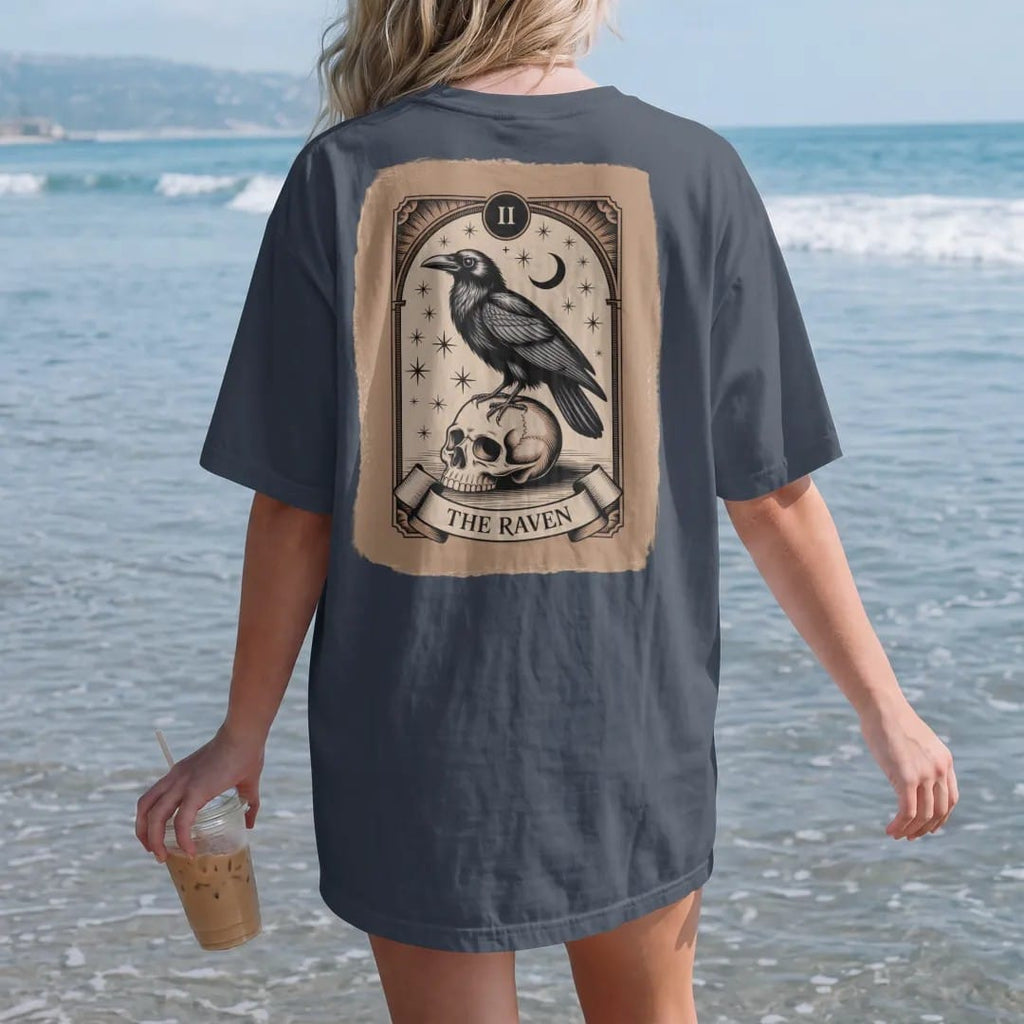 Woman on the beach in a Midnight tee, back view: back print: raven perched on a skull inside an ornate tarot-card frame with stars and a crescent moon, fine Victorian engraving style, "THE RAVEN" on a banner at the bottom in elegant high-contrast…