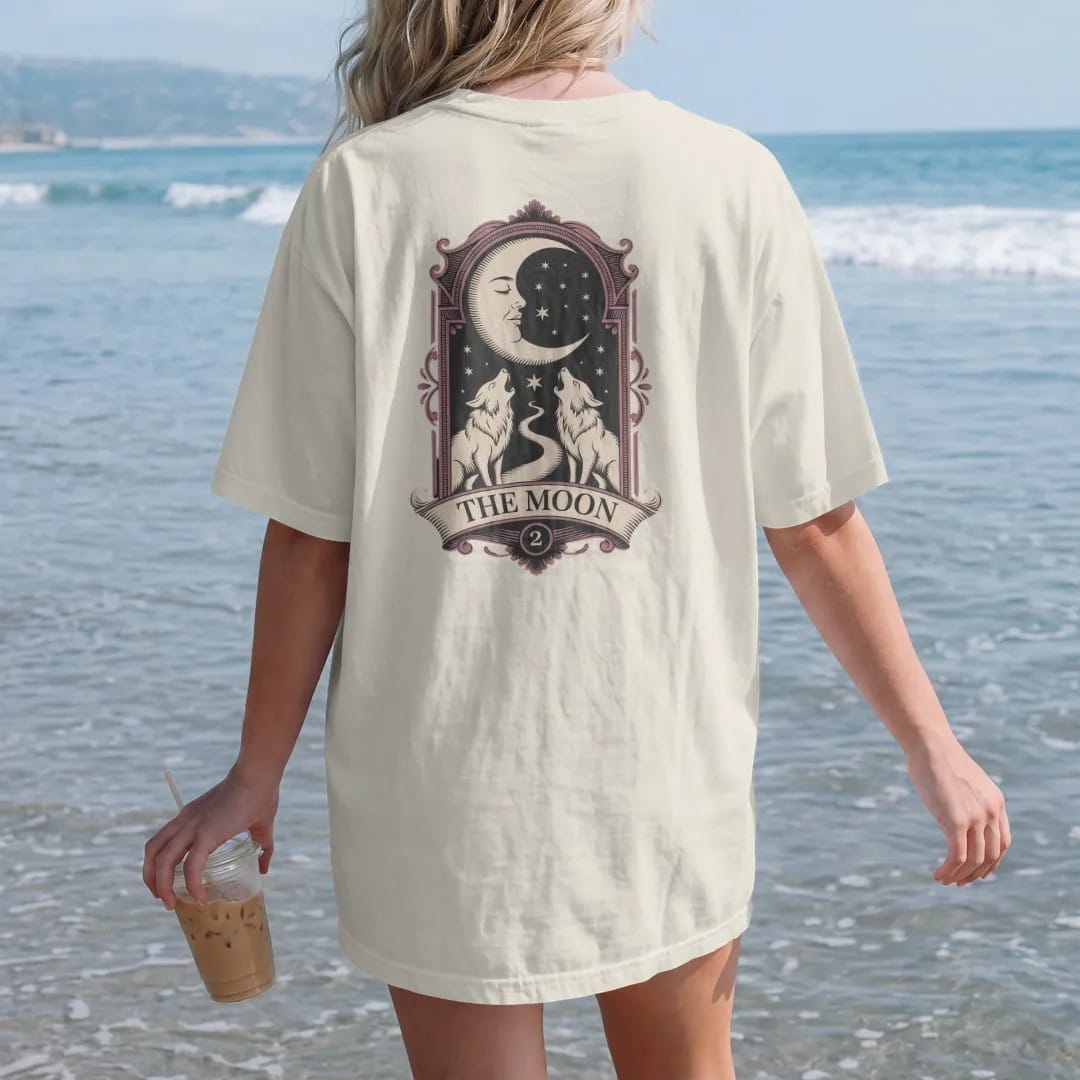Woman on the beach in a Ivory tee, back view: back print: crescent moon with a sleeping face above two howling wolves and a winding path inside an ornate tarot-card frame, detailed engraving style, "THE MOON" on a banner below in elegant…