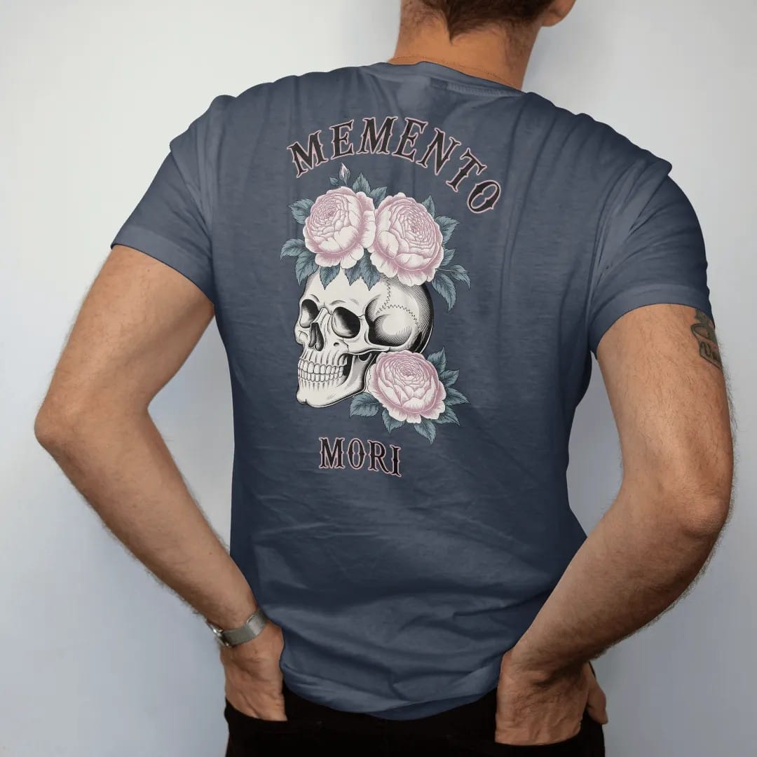 Man wearing a Midnight tee, seen from the back: back print: human skull crowned with roses, peonies and trailing ivy, fine Victorian engraving with stippled shading, "MEMENTO" above in ornate gothic blackletter, "MORI" below in small tracked…