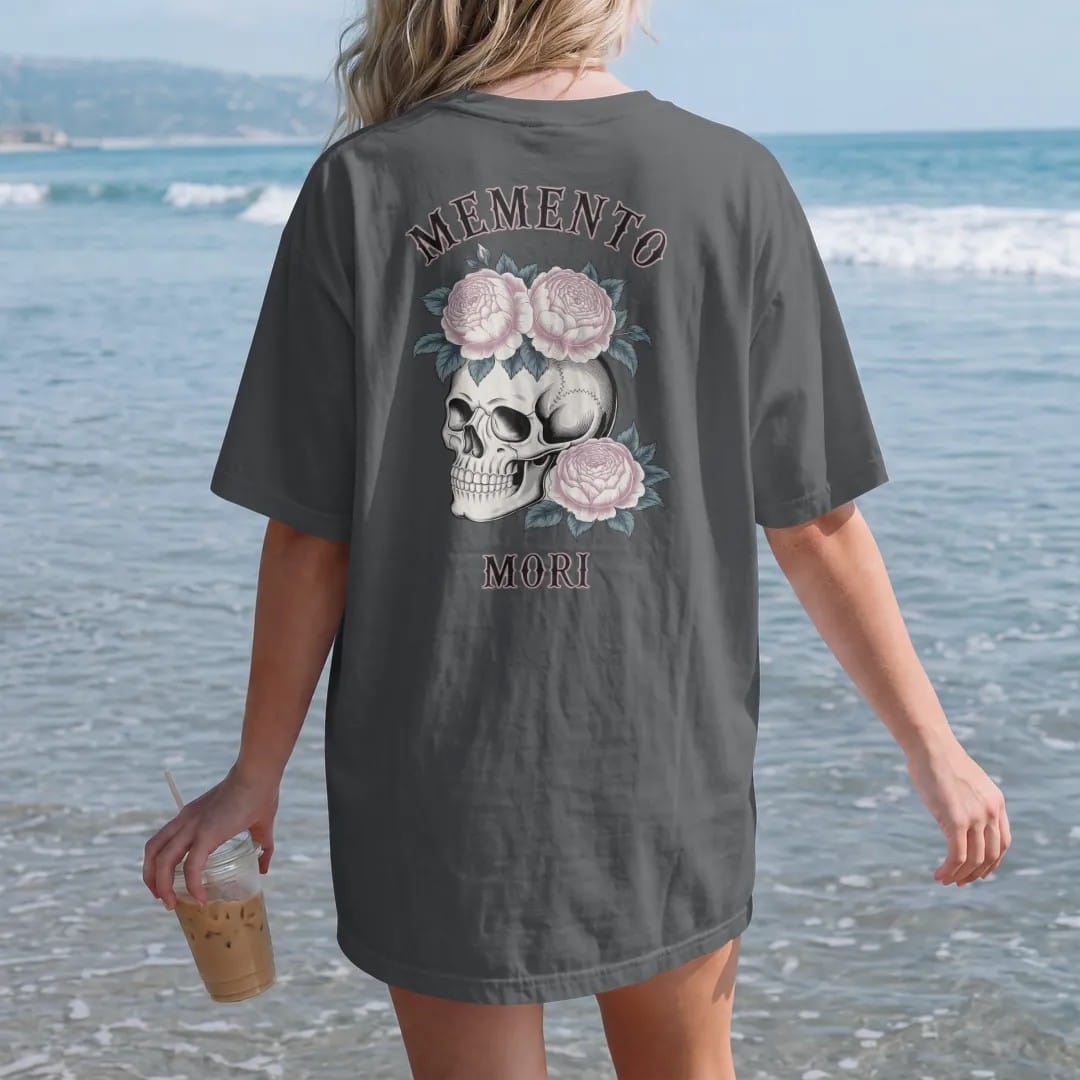 Woman on the beach in a Graphite tee, back view: back print: human skull crowned with roses, peonies and trailing ivy, fine Victorian engraving with stippled shading, "MEMENTO" above in ornate gothic blackletter, "MORI" below in small tracked…