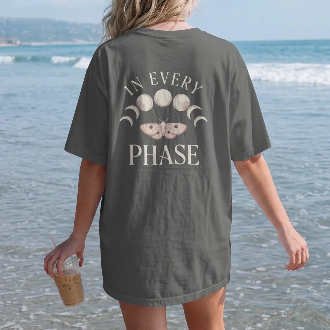 Woman on the beach in a Pepper tee, back view: back print: row of eight moon phases arched over a single small moth, fine-line engraving with stippled shadows, "IN EVERY" above in thin copperplate script, "PHASE" below in elegant high-contrast…