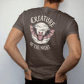 Man wearing a Espresso tee, seen from the back: back print: bat with outstretched wings in front of a full moon with scalloped cloud edges, fine engraving with crosshatching, "CREATURE" above in ornate gothic blackletter, "OF THE NIGHT" below in…