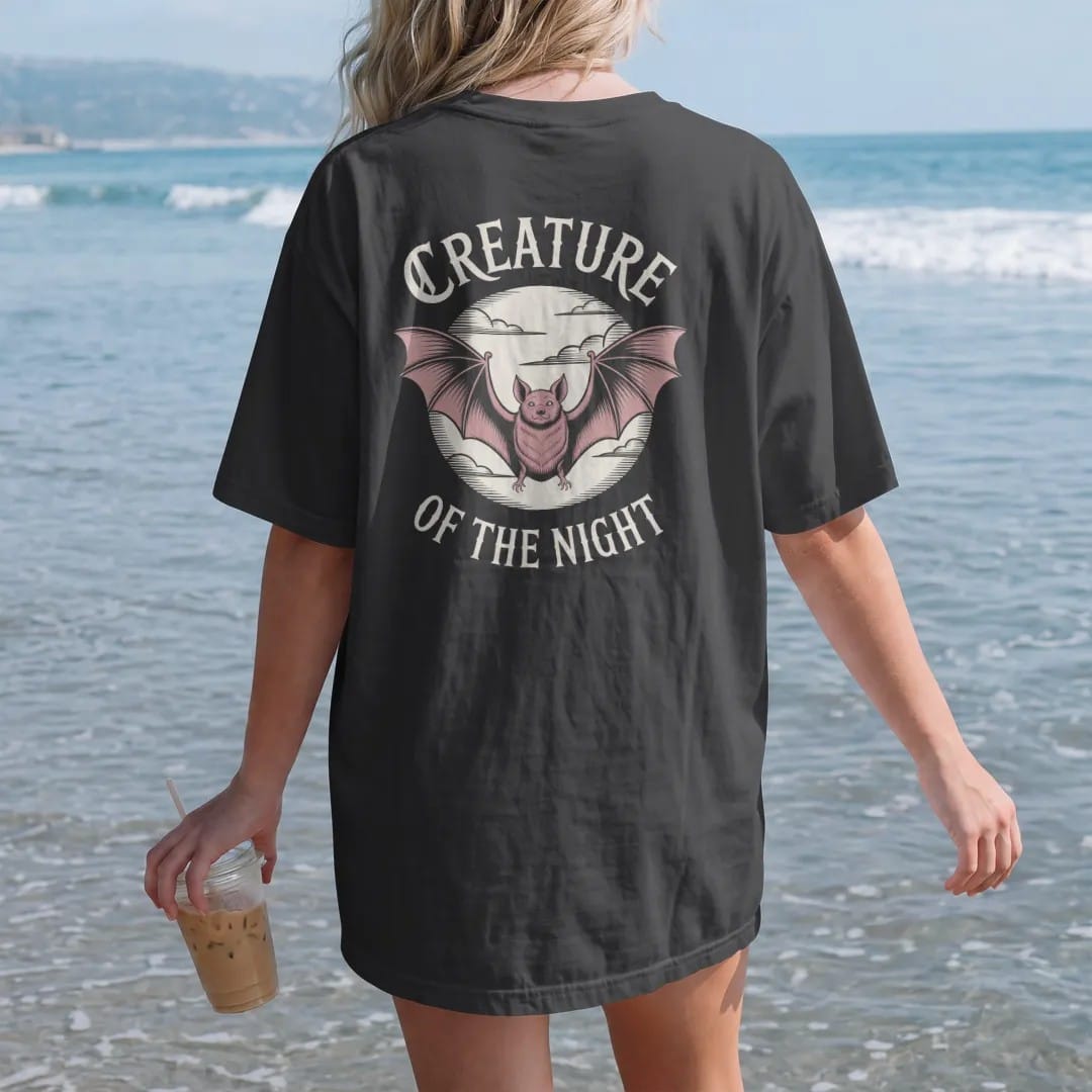 Woman on the beach in a Black tee, back view: back print: bat with outstretched wings in front of a full moon with scalloped cloud edges, fine engraving with crosshatching, "CREATURE" above in ornate gothic blackletter, "OF THE NIGHT" below in…