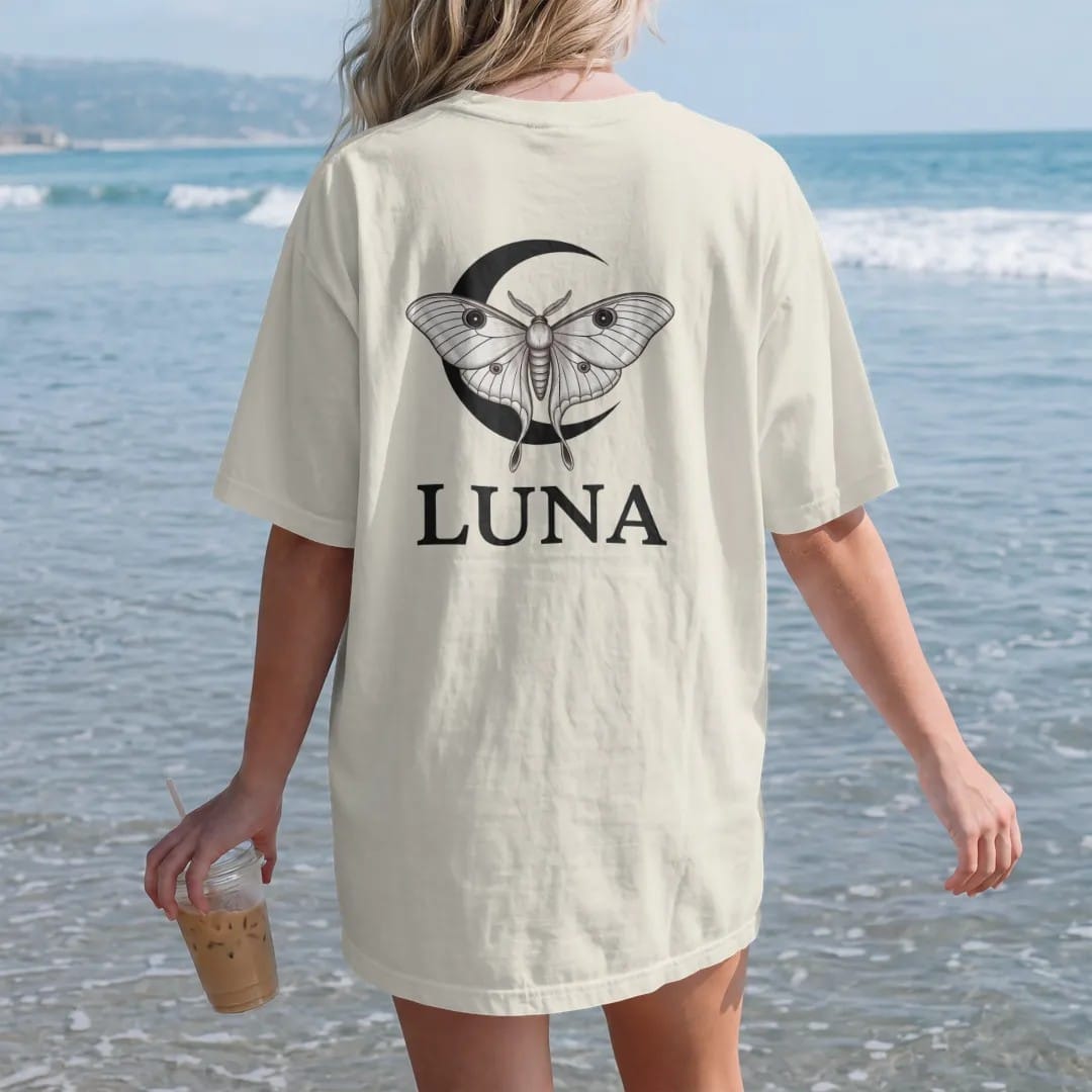 Woman on the beach in a Ivory tee, back view: back print: luna moth with long trailing wing tails over a crescent moon, delicate naturalist engraving style, "LUNA" below in elegant high-contrast serif caps with wide tracking, printed in ink black,…