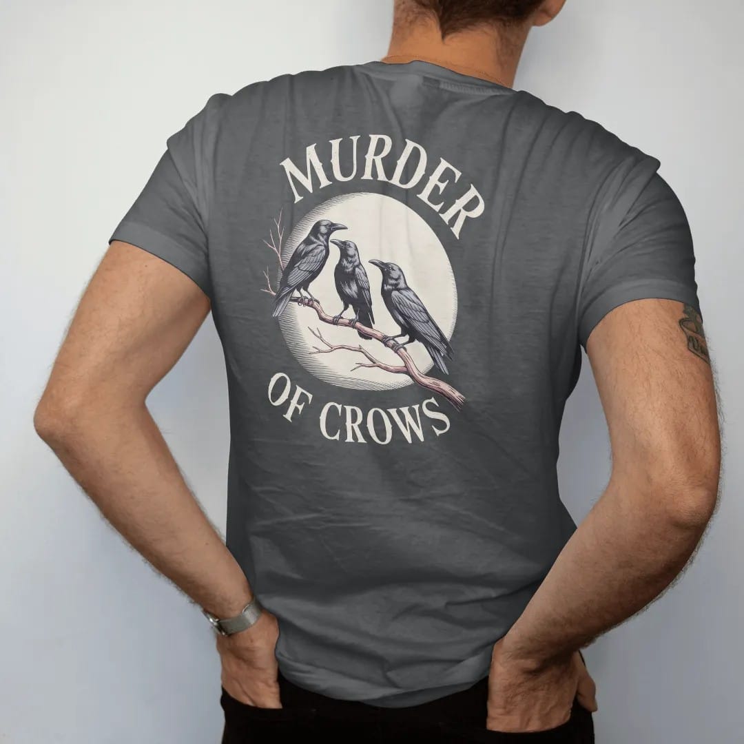 Man wearing a Graphite tee, seen from the back: back print: flock of crows perched along a bare twisted branch under a moon, fine engraving style, "MURDER" above in ornate gothic blackletter, "OF CROWS" below in small tracked engraved caps, printed…