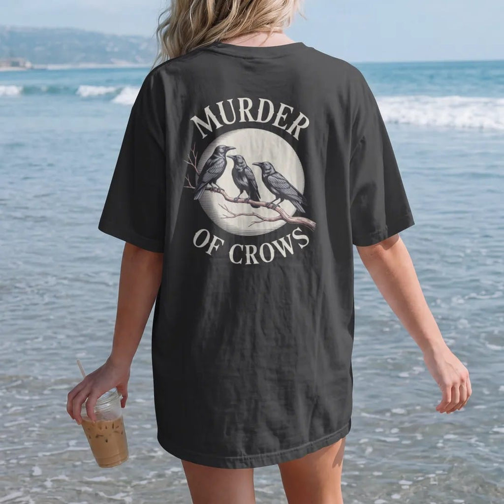 Woman on the beach in a Black tee, back view: back print: flock of crows perched along a bare twisted branch under a moon, fine engraving style, "MURDER" above in ornate gothic blackletter, "OF CROWS" below in small tracked engraved caps, printed…