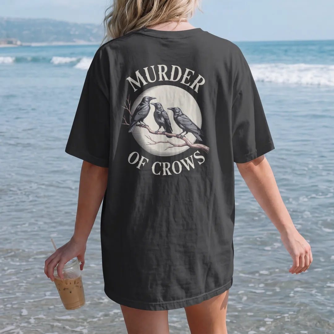 Woman on the beach in a Black tee, back view: back print: flock of crows perched along a bare twisted branch under a moon, fine engraving style, "MURDER" above in ornate gothic blackletter, "OF CROWS" below in small tracked engraved caps, printed…