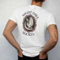Man wearing a White tee, seen from the back: back print: great horned owl with spread wings on a crescent moon inside an ornate oval frame, detailed Victorian engraving, "NIGHT OWL" arched above in elegant high-contrast serif caps, "SOCIETY" below…