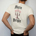 Man wearing a Ivory tee, seen from the back: back print: ornate candelabra with three dripping candles and a moth circling the flames, Victorian engraving with stippled glow, "BURN" above in ornate gothic blackletter, "BRIGHT" below in thin…